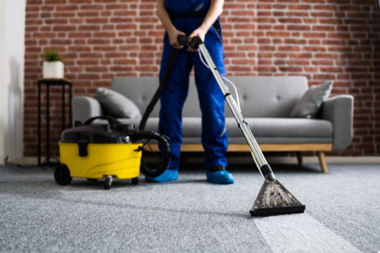 A man wearing blue overalls uses a vacuum cleaner to clean a carpet, ensuring it is free of dirt and debris.